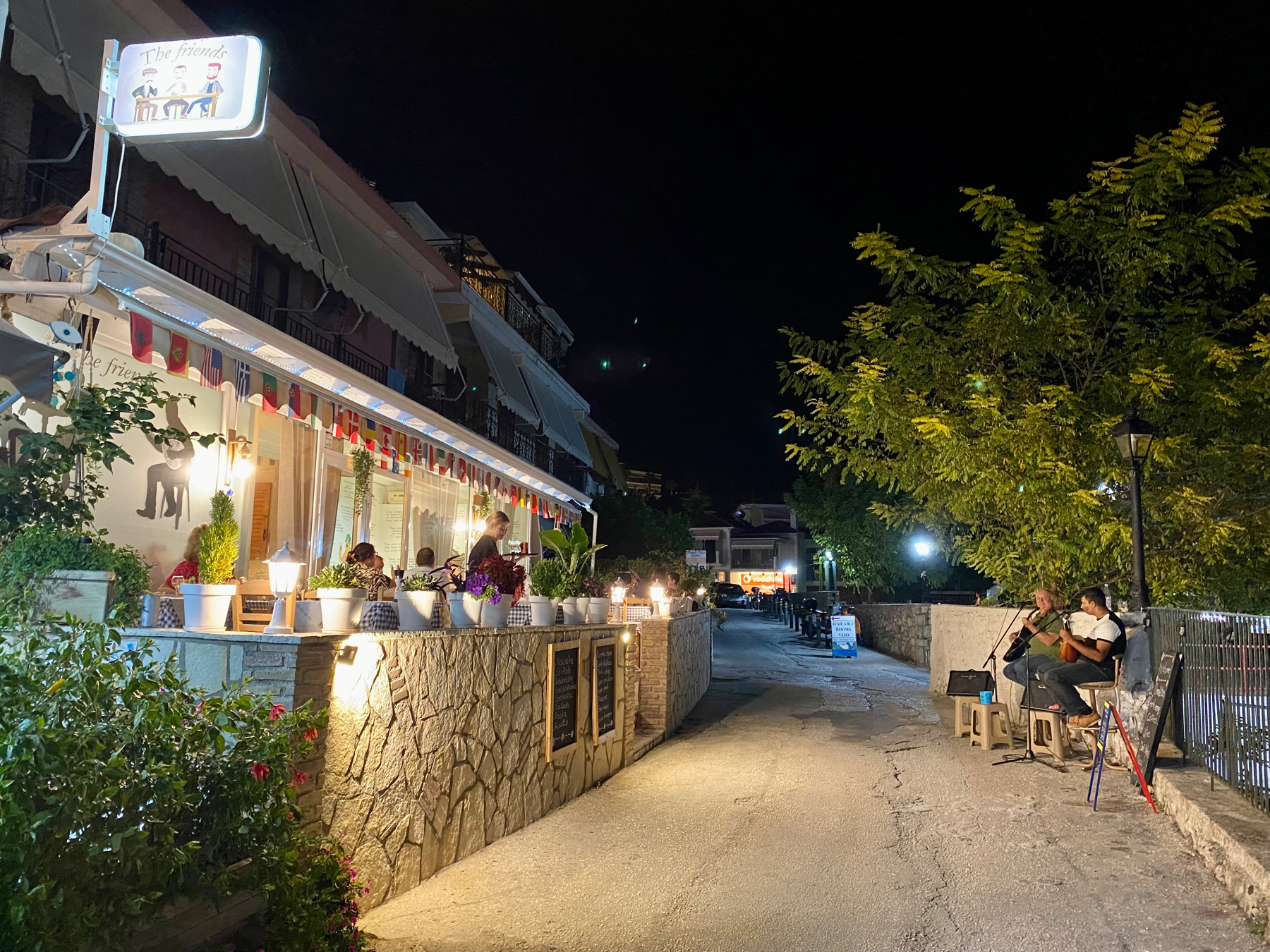 Narrow road, to the left restaurant with outdoor seating, to the right two seated musicians playing guitar and bouzouki 