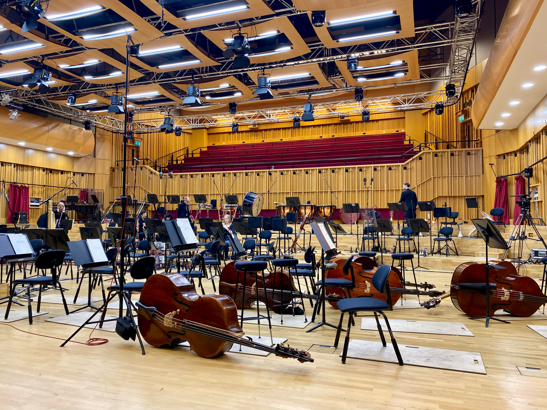 Orchestra setup with chairs, sheet music stands, and various instruments including violins, cellos, and basses on a concert hall stage with a wooden interior and modern lighting. Few musicians are present, suggesting a rehearsal break or preparation for a performance