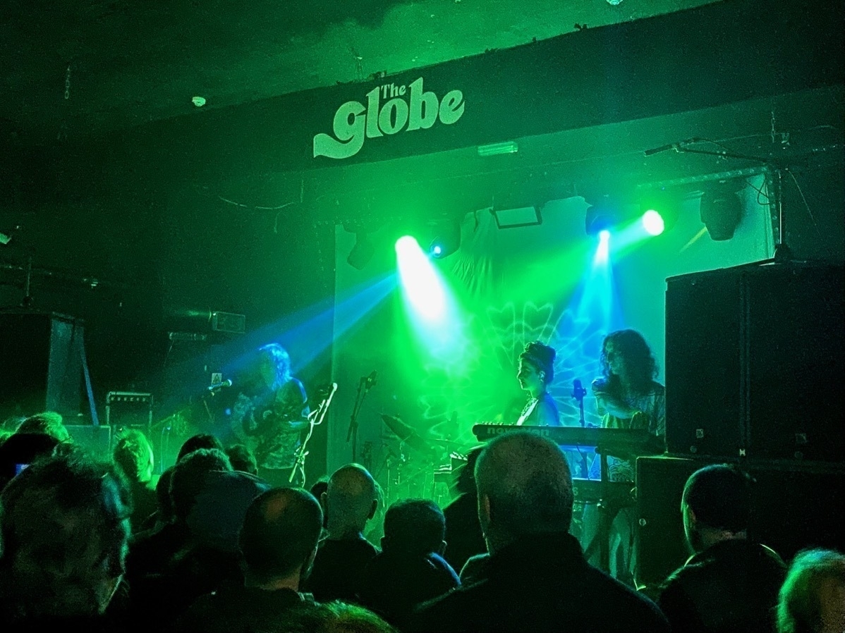 A live music performance at The Globe venue with band members illuminated by green and blue stage lights, and an audience watching the show